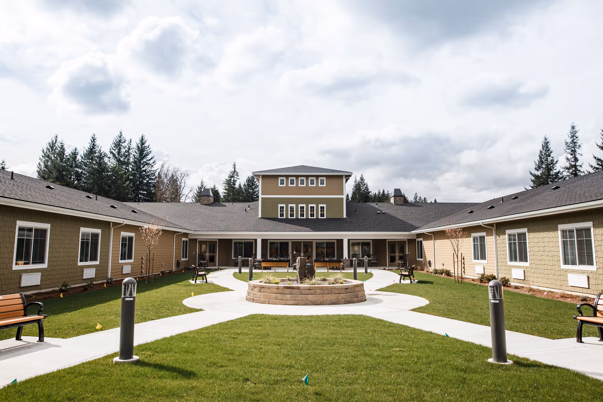 Outdoor courtyard area of a senior living facility with green grass, paved walkways, benches, and a circular raised planter in the center. The building surrounds the courtyard with multiple windows and a central two-story section. Trees and a cloudy sky are visible in the background.
