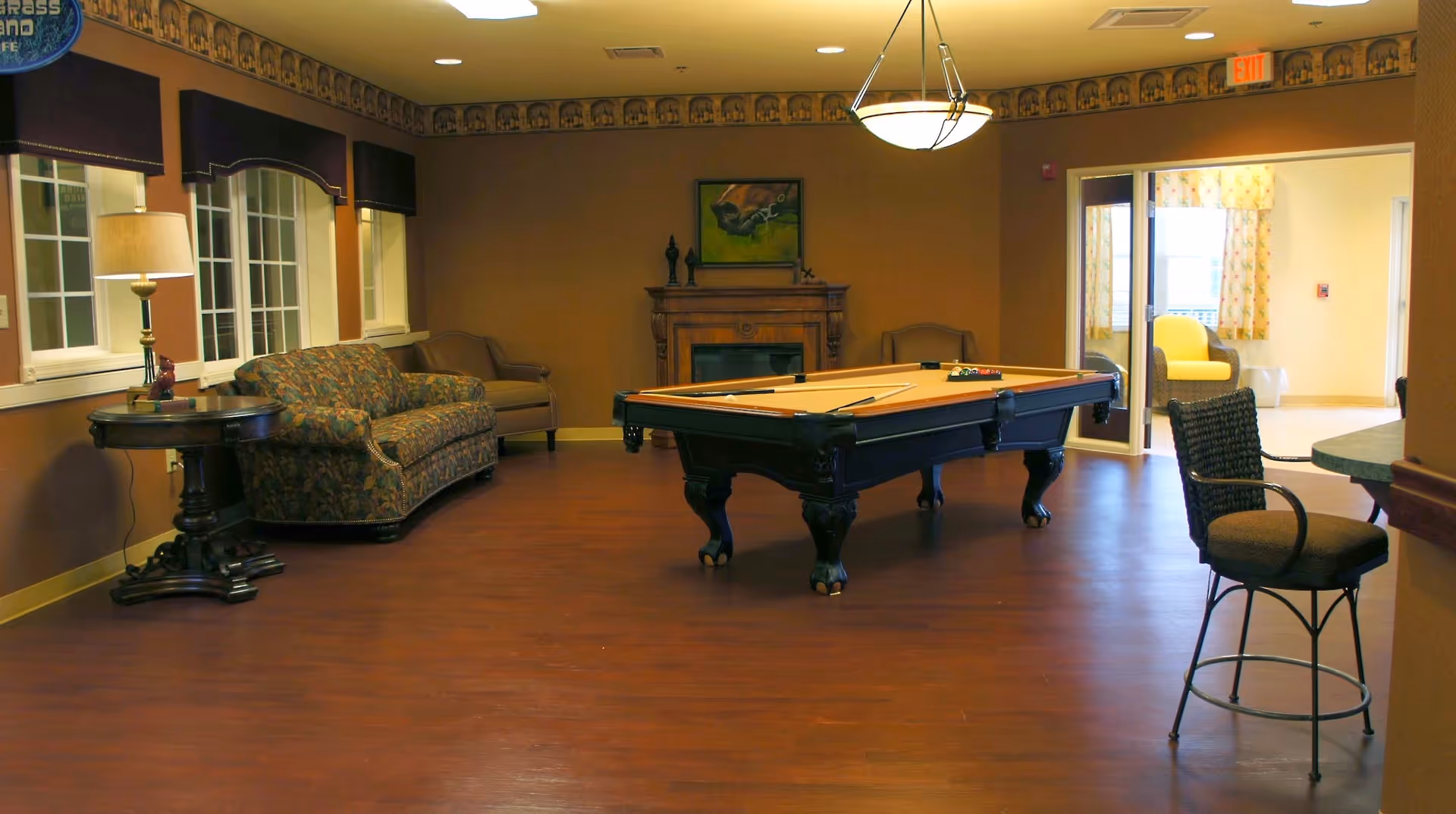 A spacious room with a pool table in the center, a floral patterned sofa and armchair along the left wall, a round side table with a lamp, and a fireplace with a painting above it on the far wall. There is a high chair near a counter on the right side and a doorway leading to a brightly lit room with yellow chairs and floral curtains.