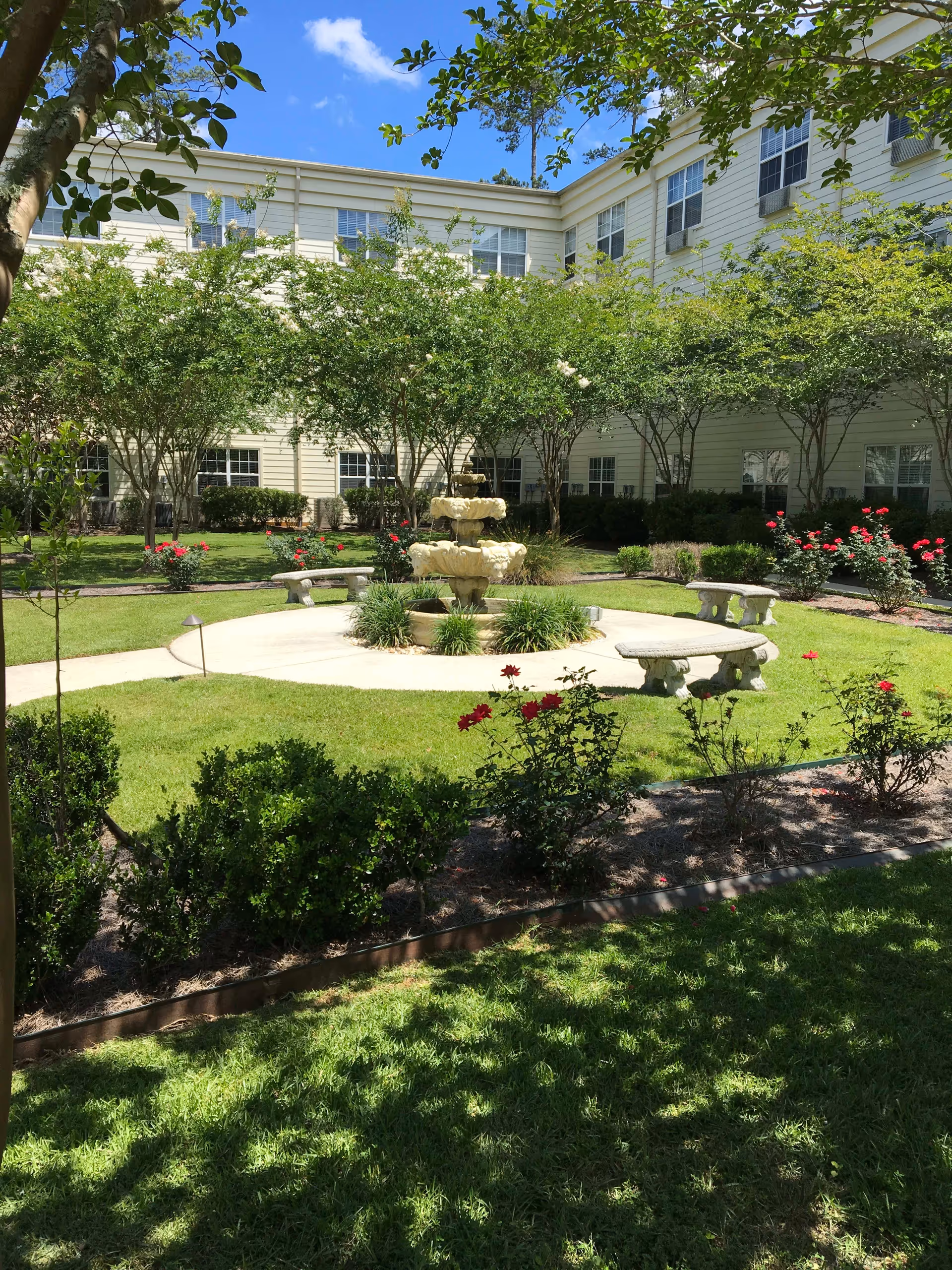 A landscaped outdoor courtyard area with a central tiered stone fountain surrounded by a circular paved walkway. There are stone benches around the fountain and well-maintained green grass, bushes, and flowering plants. The courtyard is enclosed by a multi-story building with numerous windows, and the sky is clear with a few clouds.