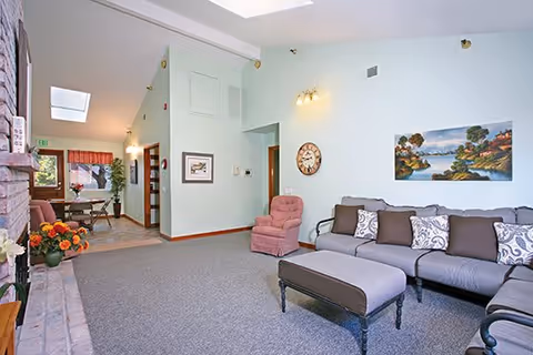A spacious living room area with a gray sectional sofa adorned with patterned and solid pillows, a matching ottoman, and a pink armchair. The walls are light blue with a landscape painting and a round clock hanging. The room has carpeted flooring and is adjacent to a dining area visible in the background with a table and chairs near windows with red valances. There is a fireplace on the left side with a flower arrangement on the hearth.