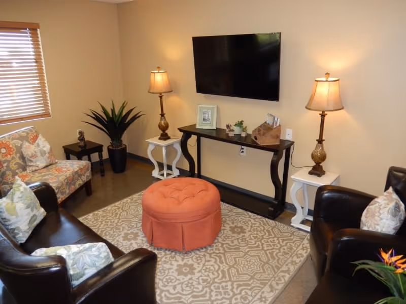 A cozy living room with two dark leather armchairs and a floral upholstered chair arranged around a patterned area rug. In the center is a round, orange tufted ottoman. Against the wall is a black console table with two matching lamps on white side tables, a flat-screen TV mounted above, and small decorative plants and picture frames on the table. A window with wooden blinds is on the left side of the room.