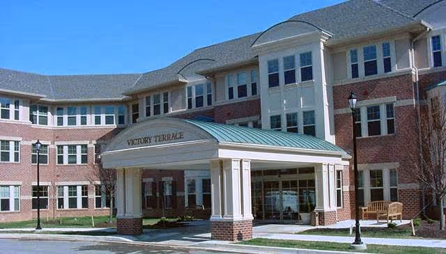 Exterior view of Victory Terrace, a multi-story senior living facility with a brick facade, numerous windows, and a covered entrance with white columns and a green roof. There are benches and street lamps near the entrance.