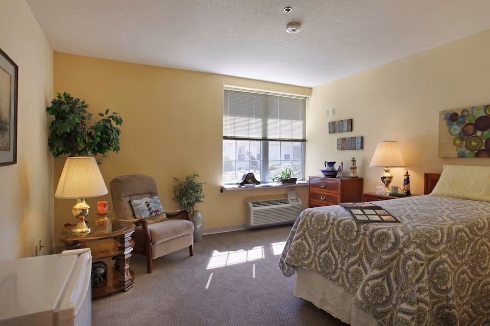 Well-lit furnished bedroom with a bed, patterned bedspread, nightstands with lamps, a recliner, and a window with blinds.