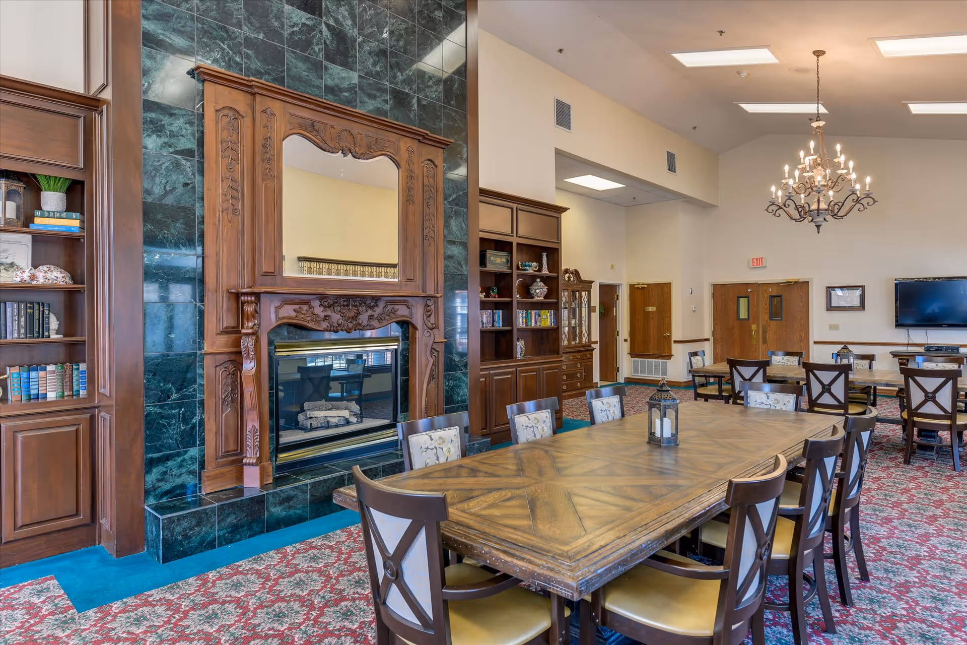 Large communal dining and activity room with a long wooden table, chandelier, ornate fireplace with a mirror, and built-in bookshelves.