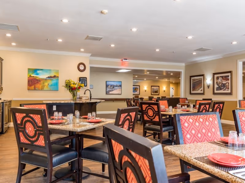 Bright communal dining room with tables and patterned chairs set with plates and glasses, artwork on the walls and a serving counter in the background.