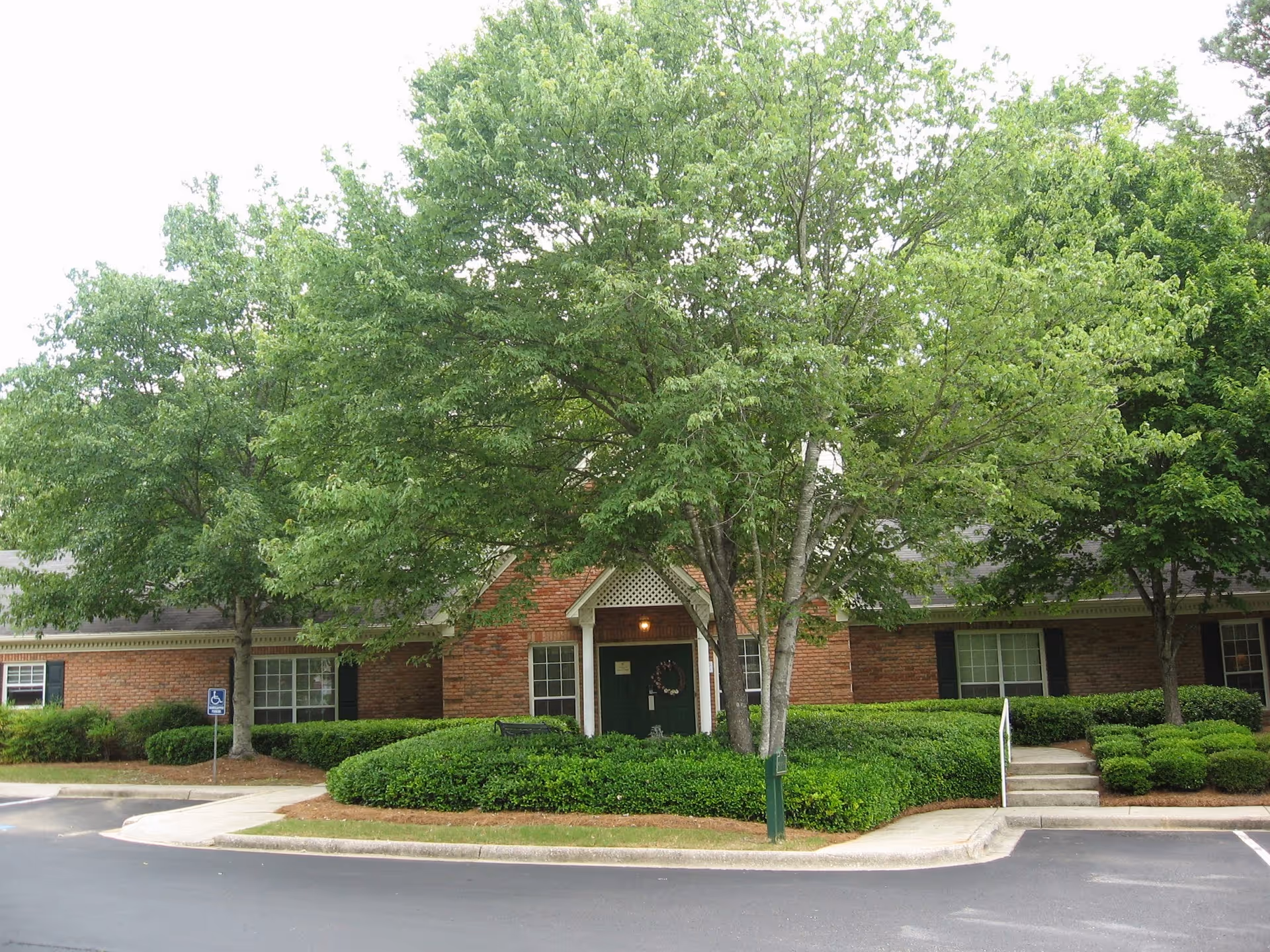 Front exterior of a single-story brick building with large trees, trimmed shrubs, a central entrance, and a parking area in front.