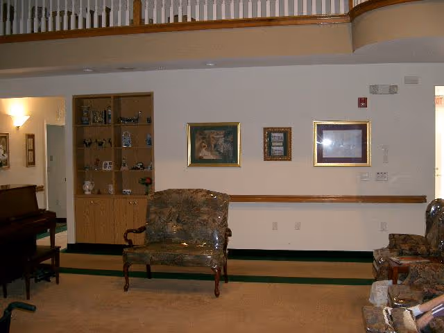 Interior view of a senior living facility common area with a floral upholstered loveseat and armchairs, a wooden piano on the left, built-in wooden shelves with decorative items, three framed pictures on the wall, and a railing above overlooking the space.