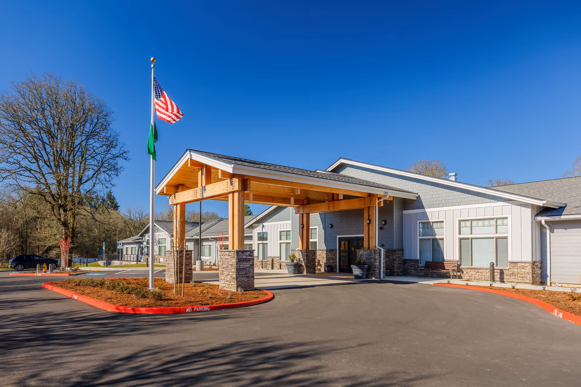 Exterior view of White Oak Village Memory Care facility showing the main entrance with a covered drop-off area supported by wooden beams and stone pillars. Two flags, including the American flag, are flying on a flagpole near the entrance. The building is surrounded by a paved driveway and landscaped areas with trees and shrubs under a clear blue sky.