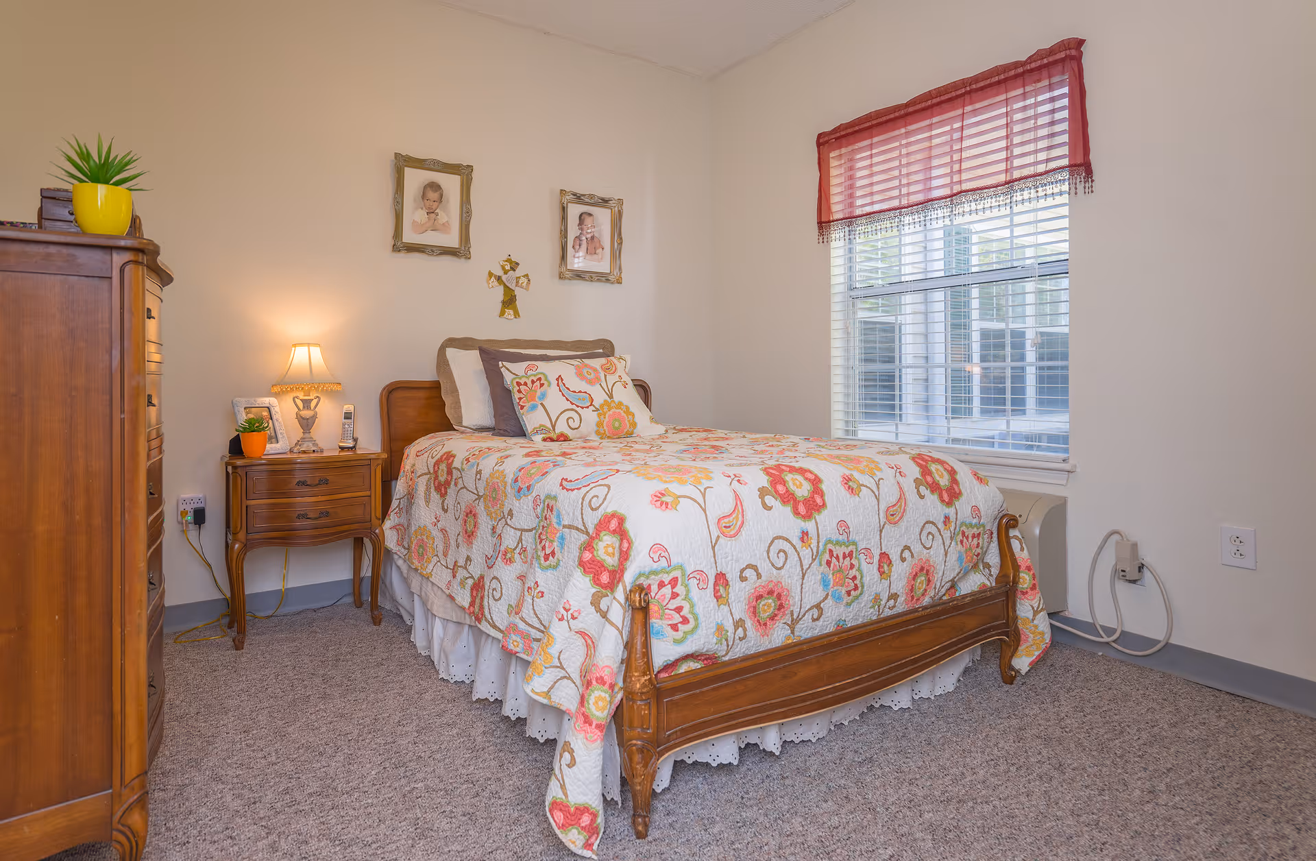 A cozy bedroom with a floral quilted bed, wooden furniture, a nightstand with a lamp, and a window with blinds.
