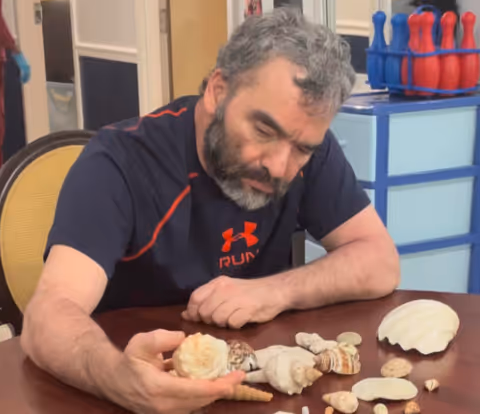 A man with gray hair and a beard is sitting at a table examining a collection of seashells laid out in front of him. Behind him, there is a blue storage unit with red and blue bowling pins on top.