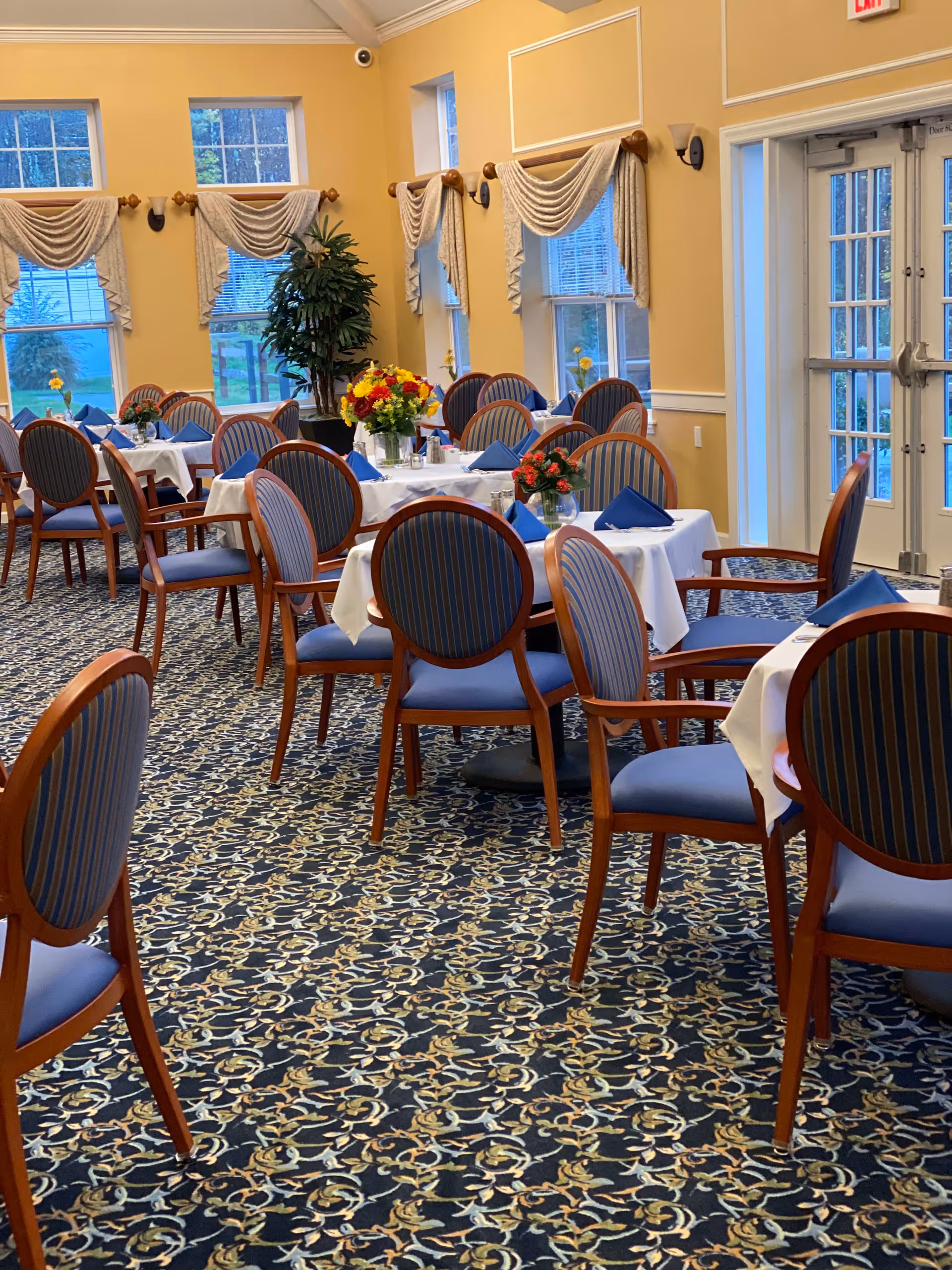 A dining room with multiple round tables covered with white tablecloths and set with blue napkins. Each table has a floral centerpiece. The room has large windows with decorative curtains, yellow walls, and patterned carpet. There are wooden chairs with blue cushioned seats and backs around the tables.
