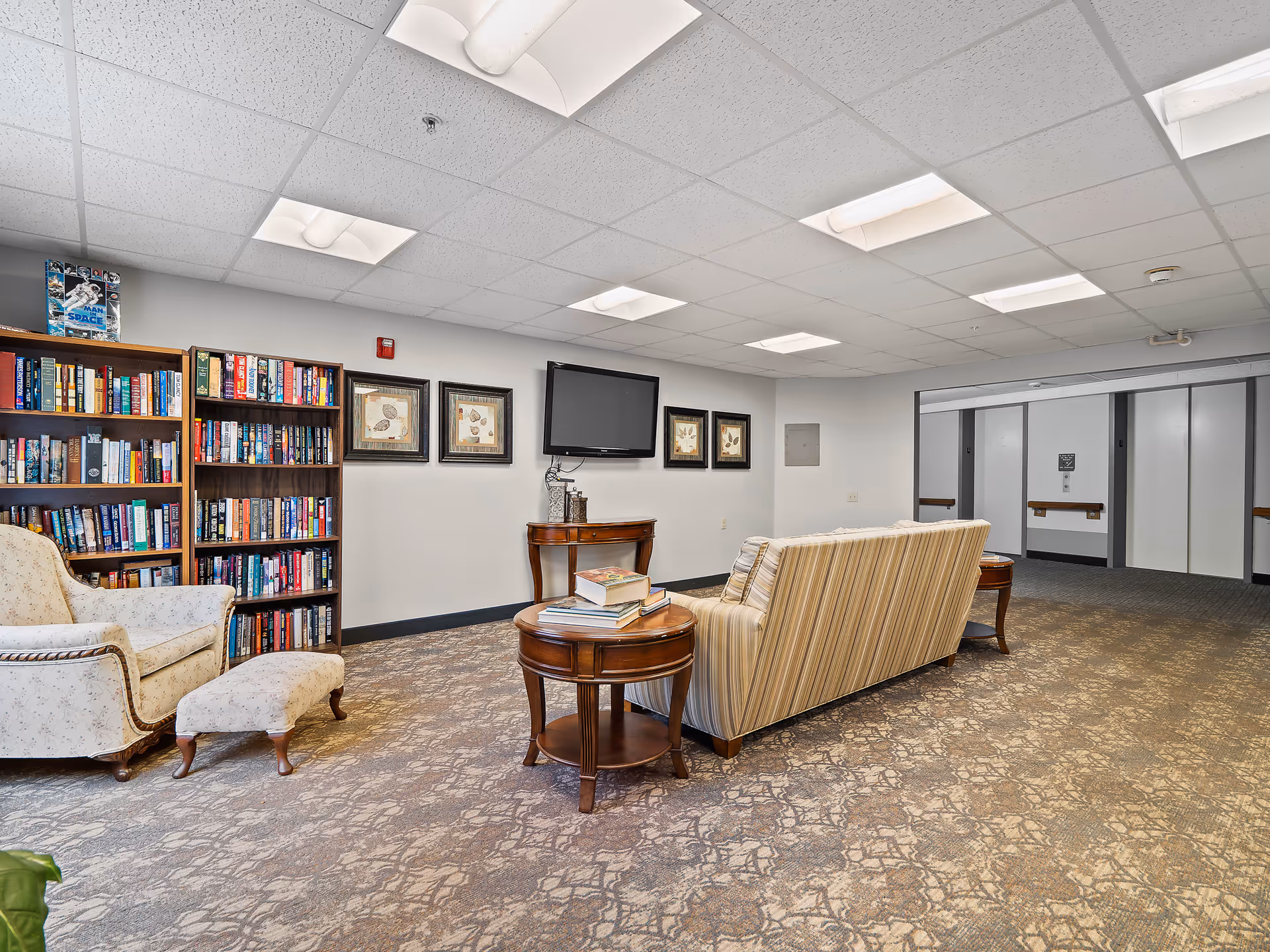 A cozy common area in a senior living facility featuring a striped sofa, a floral armchair with matching ottoman, two wooden side tables with books, and two tall bookshelves filled with books. A flat-screen TV is mounted on the wall above a small wooden console table. The room has a patterned carpet and a drop ceiling with fluorescent lights. In the background, there are elevator doors and a hallway.