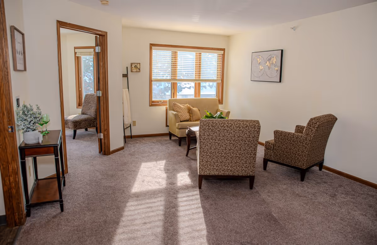 A bright living room with beige walls and carpeted floor. The room features a small beige loveseat with two pillows, two patterned armchairs, and a wooden coffee table with a plant on it. There is a large window with wooden blinds letting in natural light. A framed world map hangs on the wall. To the left, there is a small wooden side table with a plant and a green glass, and an open doorway leading to another room with a patterned chair.