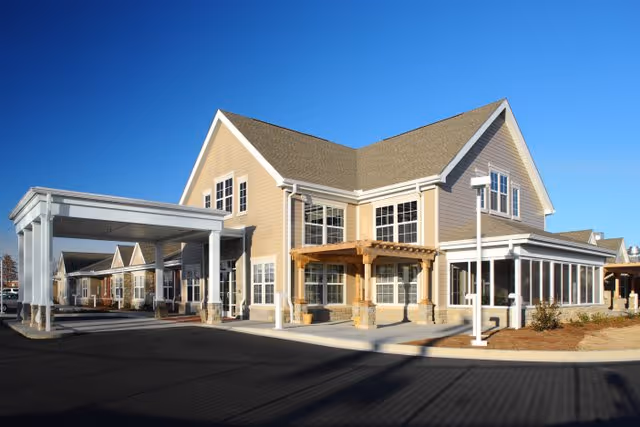 Exterior view of a senior living facility building with beige siding, white trim, and a covered entrance. The building has multiple windows and a small wooden pergola near the entrance. The sky is clear and blue.