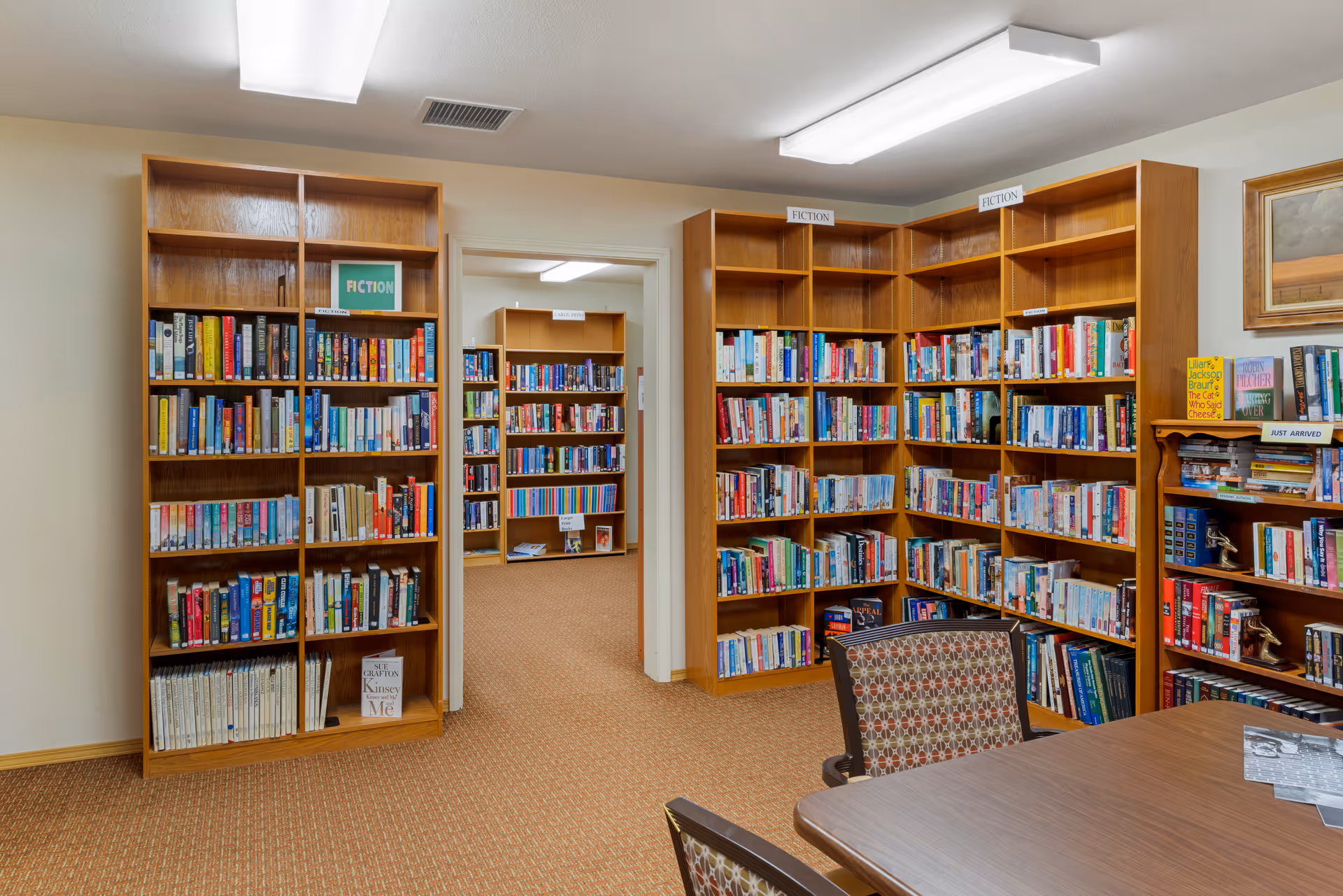 Interior view of a library room with wooden bookshelves filled with books labeled 'Fiction'. There is a wooden table with patterned chairs in the foreground and a doorway leading to another room with more bookshelves.