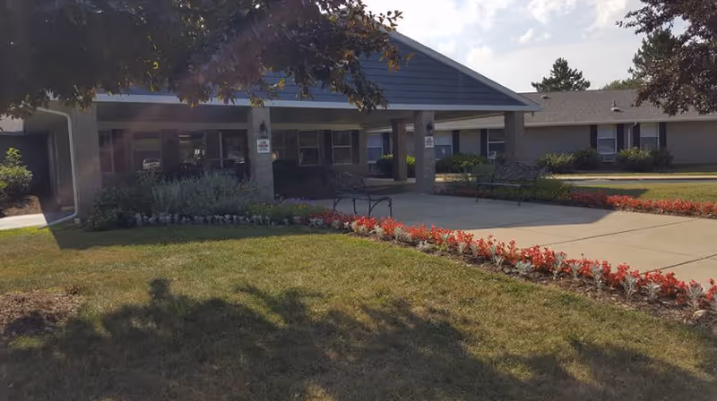 Front exterior view of a single-story building with a covered entrance, benches, and landscaped flower beds with red flowers and green grass under a partly cloudy sky.