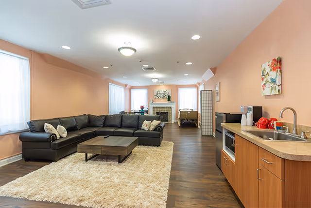 A spacious living room with peach-colored walls, a large black sectional sofa with decorative pillows, a wooden coffee table on a beige shag rug, and a kitchenette area with a sink, microwave, and coffee maker. The room has large windows with white curtains, a fireplace with a decorative piece above it, and wooden flooring.