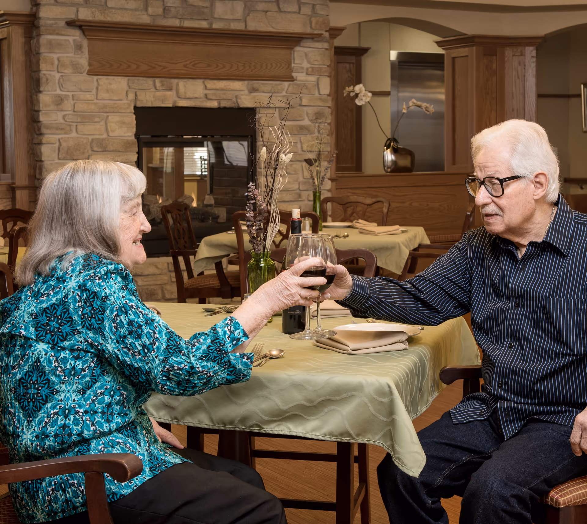 An elderly woman and an elderly man sitting at a dining table in a cozy room with a stone fireplace in the background. They are clinking wine glasses and smiling at each other. The table is covered with a green tablecloth and set with napkins and silverware.