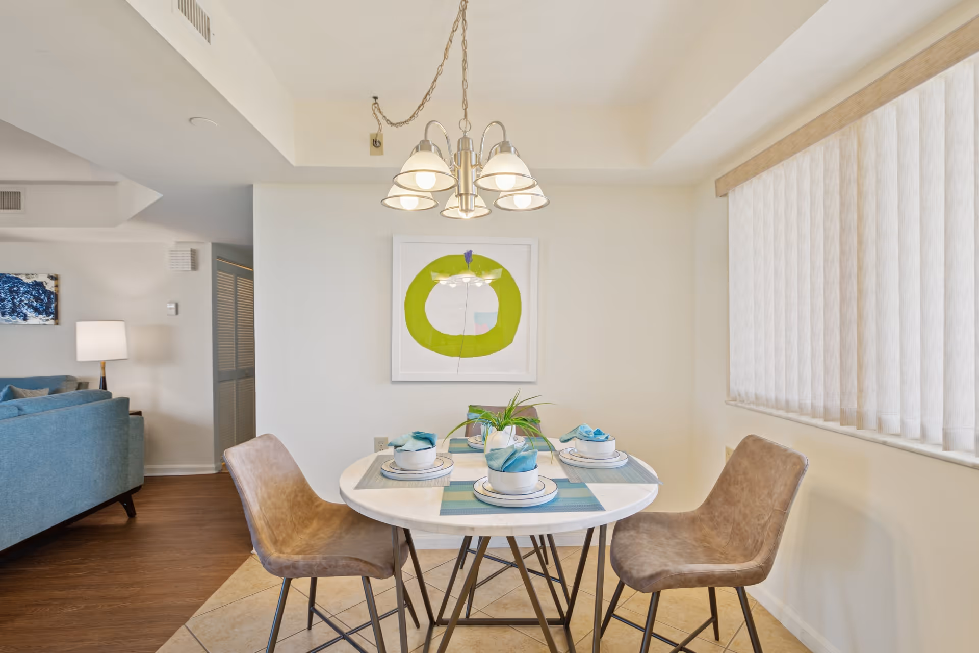 A small dining area with a round white table set for four, featuring plates, bowls, and blue napkins. Four brown cushioned chairs surround the table. A modern chandelier with five lights hangs above the table. On the wall behind the table is a framed abstract artwork with a green circular shape. To the right, there is a window with vertical blinds. Part of a living room with a blue sofa and a lamp is visible to the left.