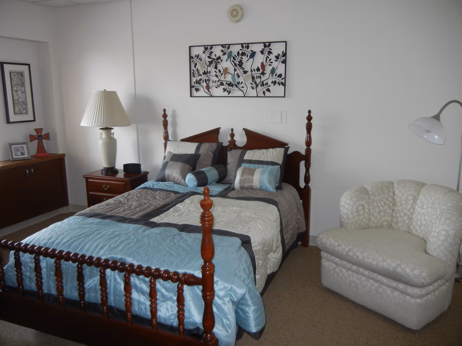 A bedroom with a wooden bed frame and a bed made with a blue, gray, and beige comforter and matching pillows. To the left of the bed is a wooden nightstand with a white lamp, a small black clock, and a cabinet with framed pictures and a red cross decoration. On the right side of the bed is a white patterned armchair and a white floor lamp. A framed artwork featuring birds on branches hangs on the wall above the bed.