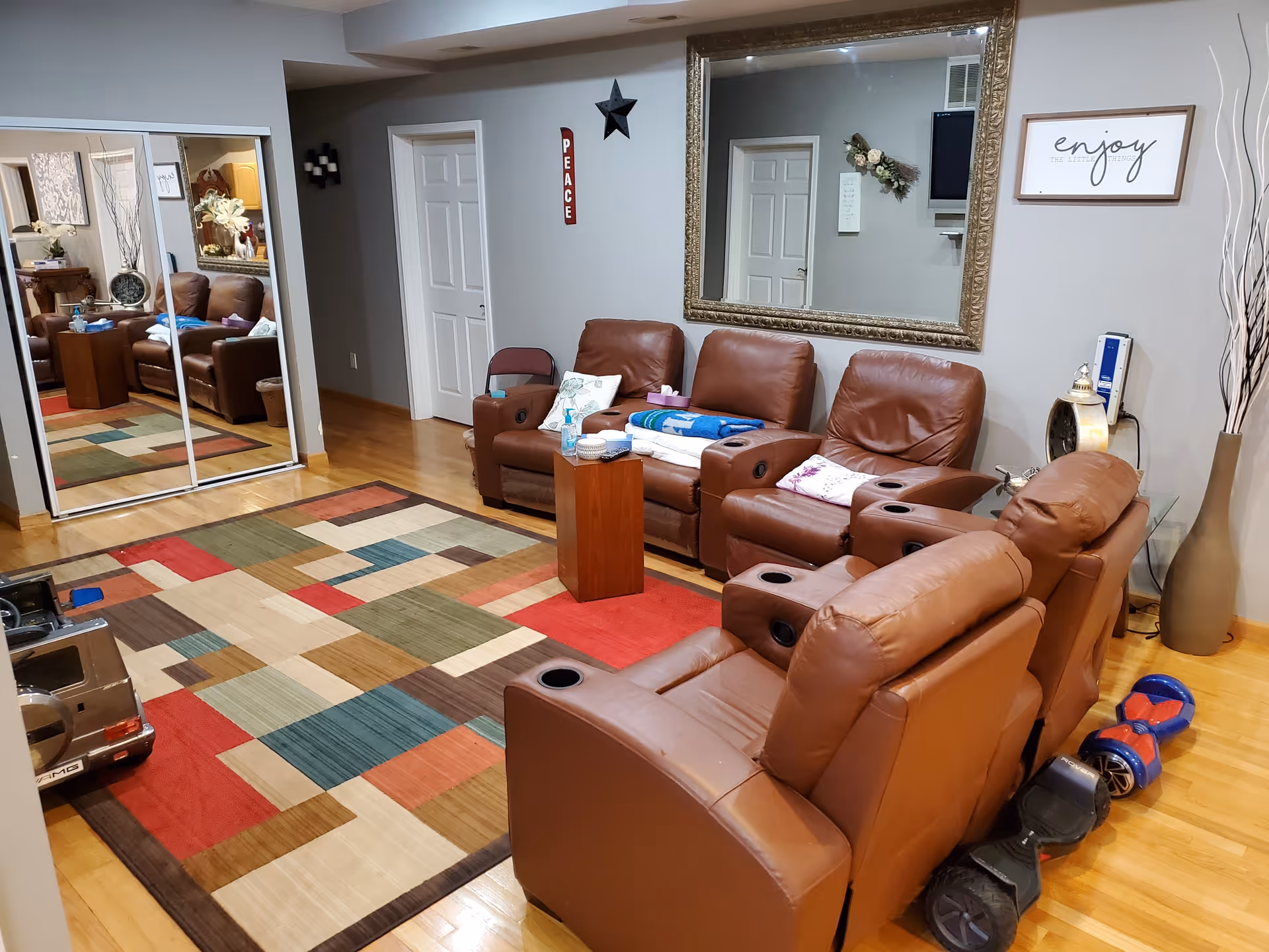 A cozy living room with five brown leather recliner chairs arranged in an L-shape around a colorful geometric patterned rug. There is a large ornate mirror on the wall above the chairs, a small wooden side table with items on it, and a framed sign that says 'enjoy'. A mirrored closet door reflects part of the room. The floor is wooden, and there are two hoverboards on the floor near the chairs.