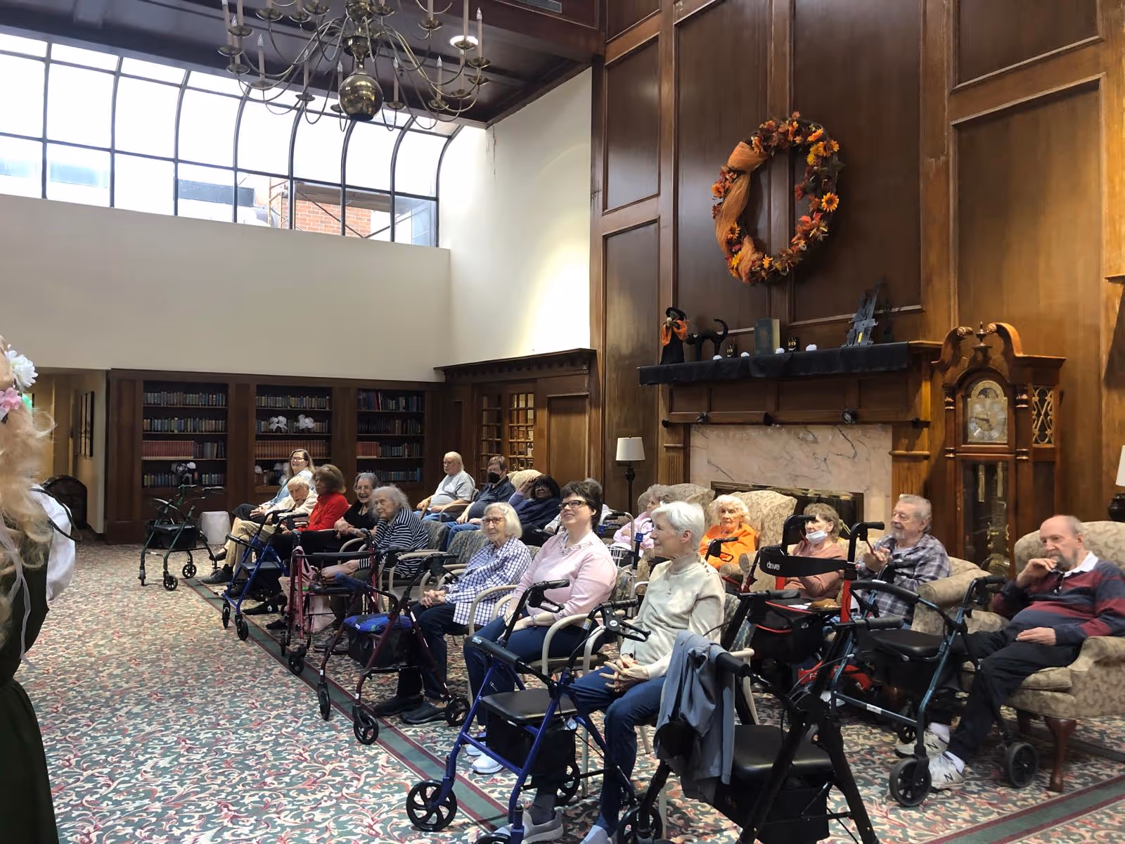 A group of elderly residents seated in a large, wood-paneled common room with a high ceiling and large windows. Many residents are using walkers and are seated in chairs arranged in rows facing the left side of the image. The room features a fireplace with a decorative wreath above it, a grandfather clock, and bookshelves along the back wall.