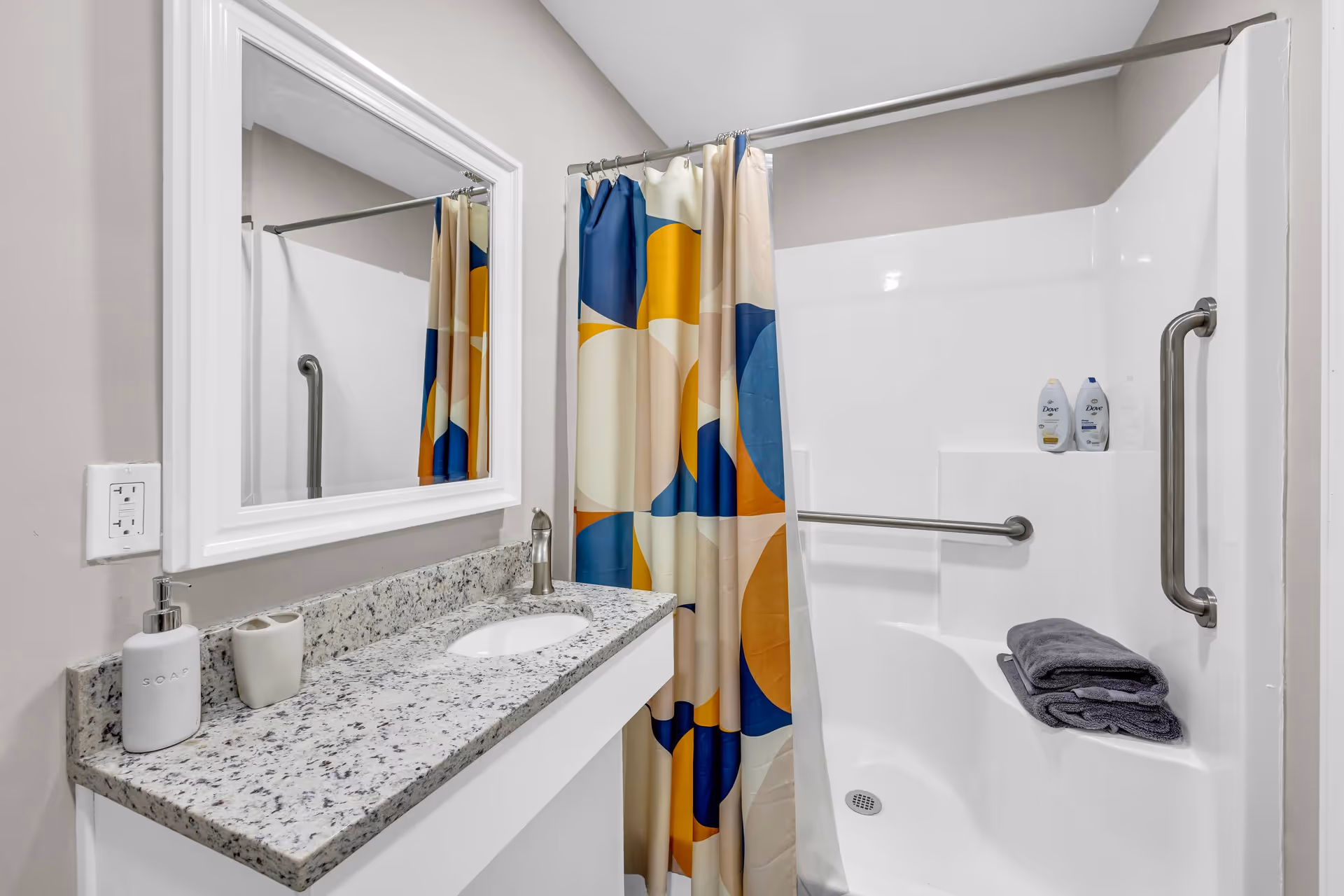 A clean and modern bathroom featuring a granite countertop with a built-in sink, a soap dispenser, and a toothbrush holder. There is a large white-framed mirror above the sink. Next to the countertop is a white shower area with a colorful shower curtain in blue, yellow, and beige tones. The shower has grab bars for safety, two bottles of Dove body wash on a built-in shelf, and two folded dark gray towels.