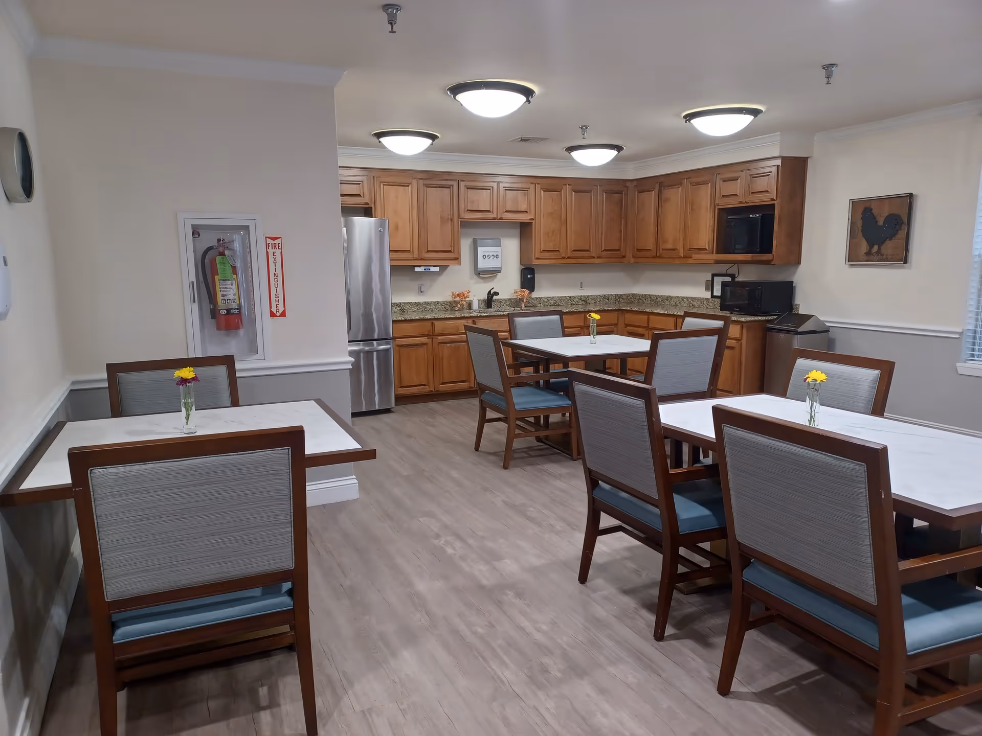 A dining area with several tables and chairs, each table decorated with a small vase containing a yellow flower. The room features wooden cabinets, a stainless steel refrigerator, a microwave, and granite countertops. The walls are light-colored with a framed picture of a rooster and a fire extinguisher mounted on one wall.