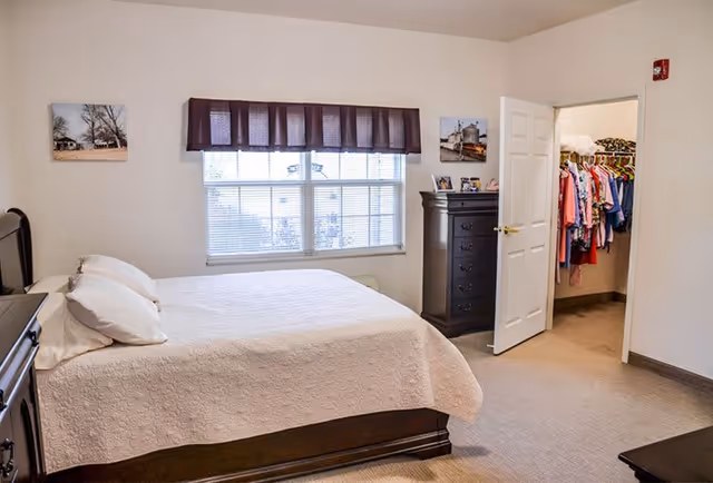 A bedroom with a large bed covered in a white quilt and two pillows. There is a window with blinds and a dark valance above it. On the right side, there is a dark wooden dresser and an open door leading to a closet filled with hanging clothes. The walls are light-colored and there are two framed pictures hanging on the wall.