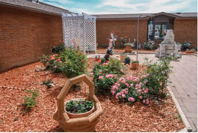A garden area with various flowering plants and shrubs surrounded by mulch. There is a wooden basket planter in the foreground, a white lattice structure against a brick wall, and a paved walkway on the right side. The sky is partly cloudy.