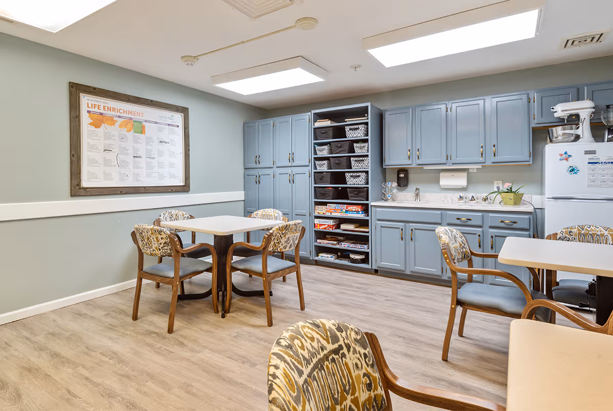 A bright room with light blue cabinets, a white refrigerator, and a countertop with a sink. There are two tables with four chairs each, featuring patterned upholstery. A bulletin board on the wall displays a 'Life Enrichment' schedule. Shelves hold board games and storage baskets. The floor is light wood, and the walls are painted a soft green with white trim.