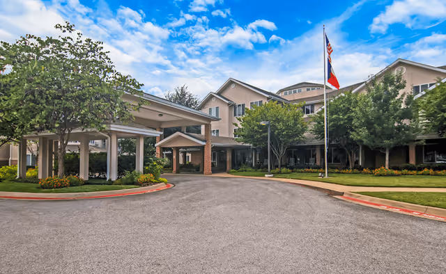 Front exterior view of Whiterock Court - Sky Active Living facility with a covered entrance, landscaped greenery, and an American flag on a flagpole under a partly cloudy blue sky.