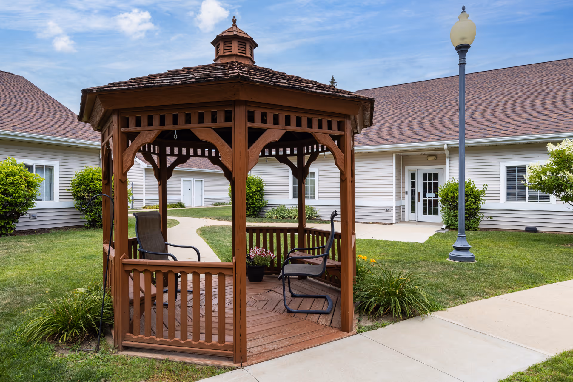Wooden gazebo with chairs in a grassy courtyard in front of a single-story light-colored building.