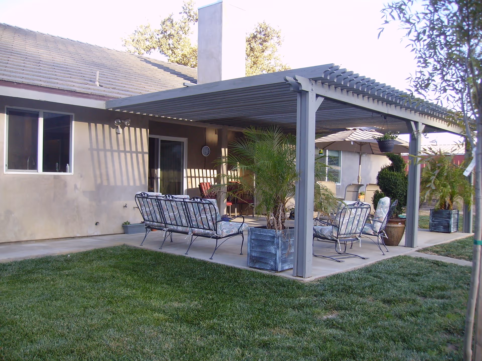Outdoor patio area with metal framed chairs and cushions under a pergola attached to a building. There are potted plants around the patio and a grassy lawn in the foreground.