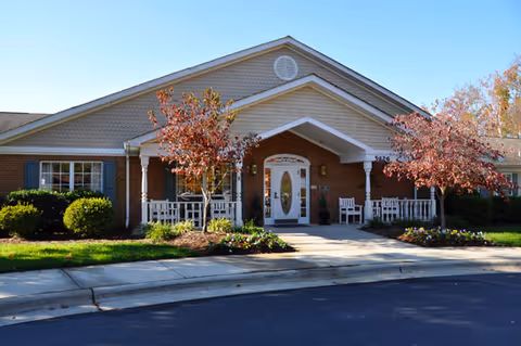 Front exterior view of a single-story brick building with a gabled roof, white trim, and a covered entrance with white columns. There are two small trees with red leaves and landscaped bushes and flowers in front of the building. The building has windows with green shutters and a paved driveway in front.