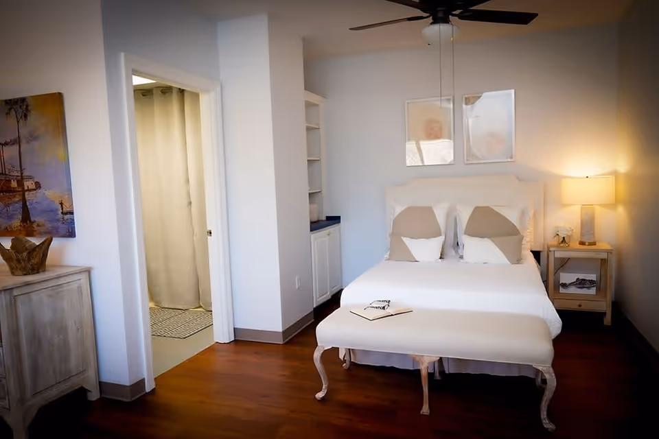 A cozy bedroom with a white bed featuring beige and white pillows, a cushioned bench at the foot of the bed, a nightstand with a lamp and decorative items, a ceiling fan, and an open doorway leading to a bathroom with a shower curtain. The room has wooden flooring, light-colored walls, and framed artwork above the bed.