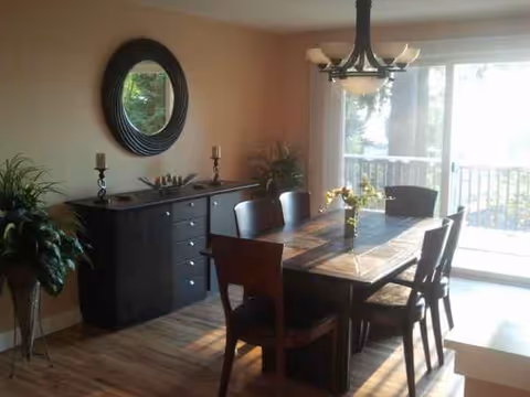 A dining room with a wooden table, chairs, plants, a sideboard, and a sliding glass door.