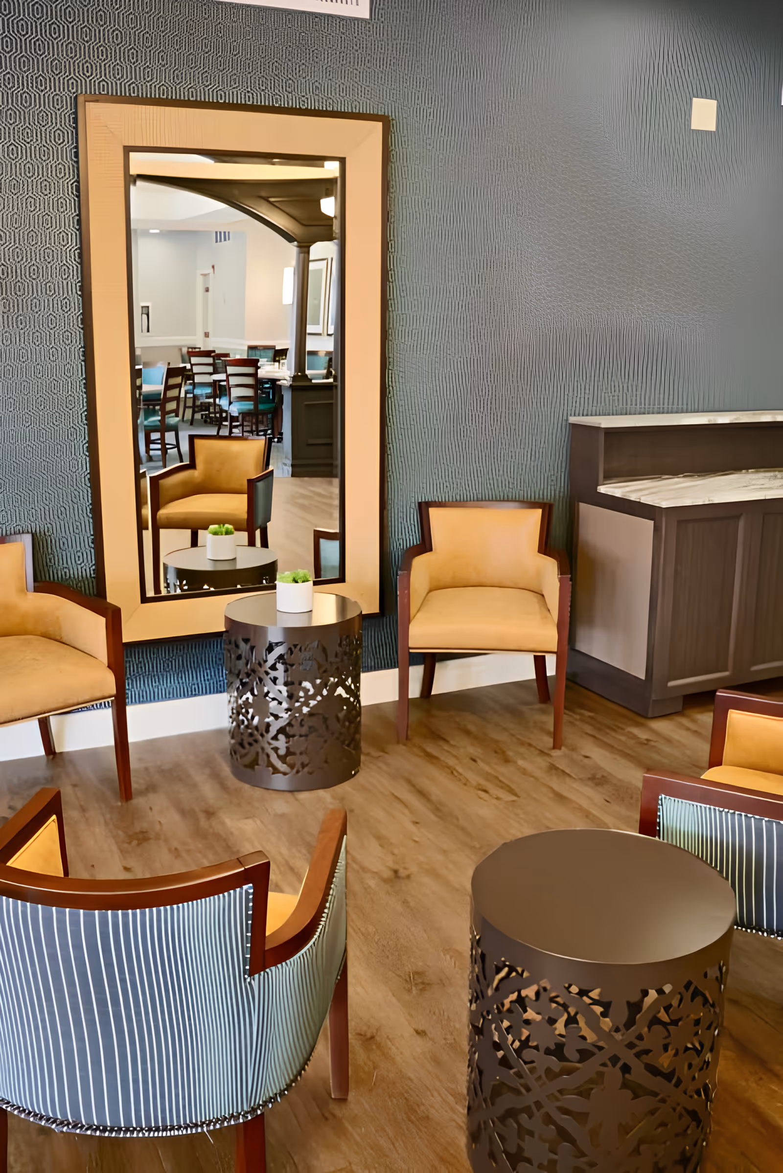 Contemporary senior living lounge with upholstered chairs, decorative metal side tables, and a large mirror reflecting a dining area.