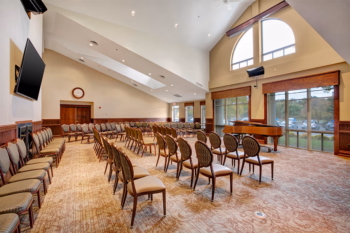 A large meeting or event room with rows of chairs arranged in a semi-circle facing a grand piano near tall windows. The room has high ceilings with recessed lighting, a wall-mounted TV, a clock above double wooden doors, and large arched windows letting in natural light with a view of parked cars and trees outside.