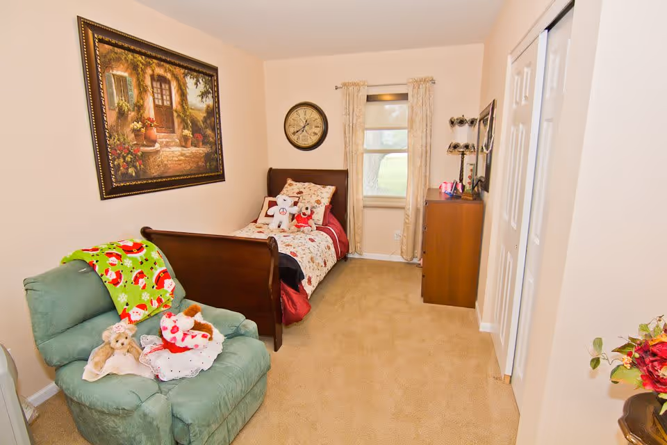A cozy bedroom with a single wooden bed adorned with floral bedding and stuffed animals. A green recliner chair with a festive blanket and more stuffed animals sits to the left. A framed painting of a rustic doorway hangs on the wall above the bed. A round clock is mounted on the wall above the bed's headboard. A window with beige curtains lets in natural light. A wooden dresser with a mirror and decorative items is positioned against the right wall. The room has beige carpet and light-colored walls.