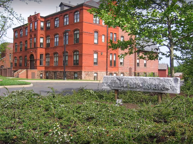 A large red brick building with multiple windows and a stone foundation, surrounded by greenery and trees. In the foreground, there is a stone sign that reads 'The Clare Estate'.