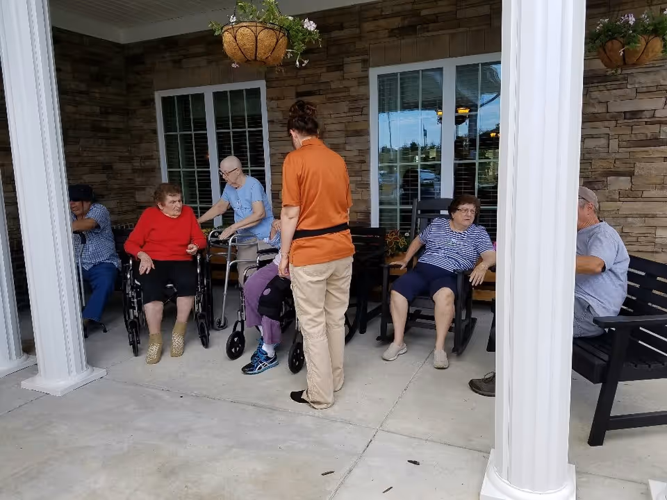 A group of elderly people sitting and standing on a covered patio area outside a building with stone walls and large windows. Some are using wheelchairs and walkers, and a caregiver in an orange shirt and beige pants is standing among them. Hanging flower baskets are visible above.