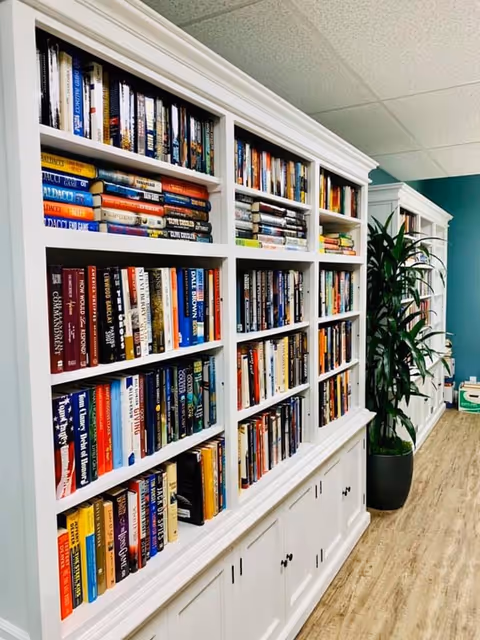 Long white built-in bookshelf filled with books along a hallway with wood flooring and a potted plant.