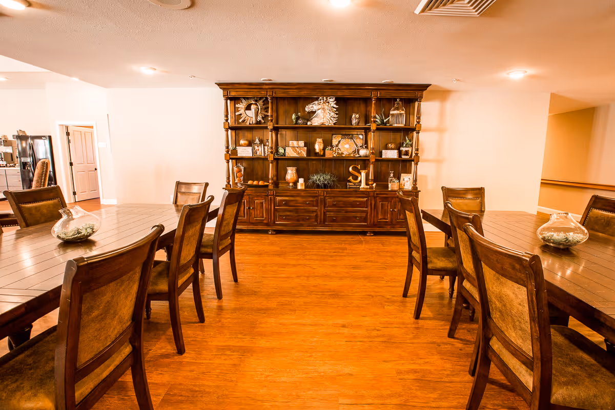 A warm, inviting dining room with two large wooden tables surrounded by chairs. A large wooden cabinet with decorative items such as vases, plants, and sculptures is against the far wall. The room has hardwood flooring and soft lighting from ceiling fixtures.