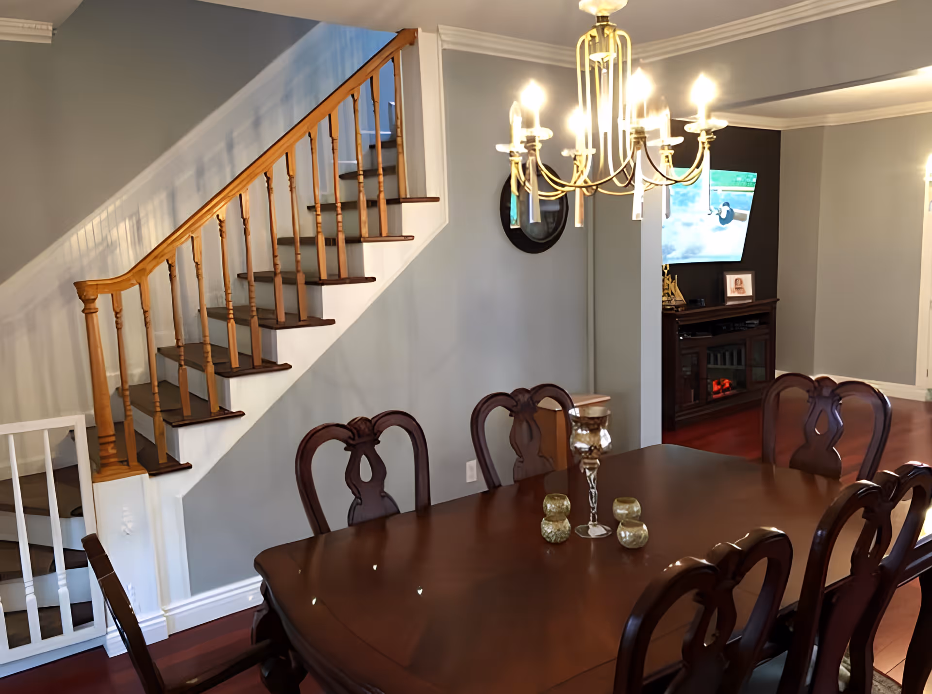 Interior view of a dining room with a wooden dining table and six chairs. A chandelier with lit bulbs hangs above the table. In the background, there is a staircase with wooden railings and a TV mounted on the wall above a dark wooden cabinet.