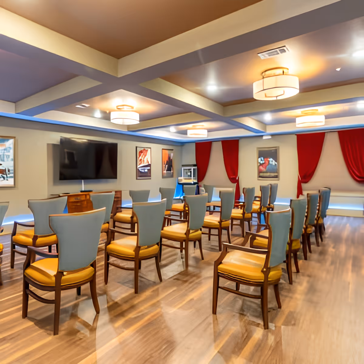 An interior activity room with rows of upholstered chairs facing a large wall-mounted TV and red-draped windows.