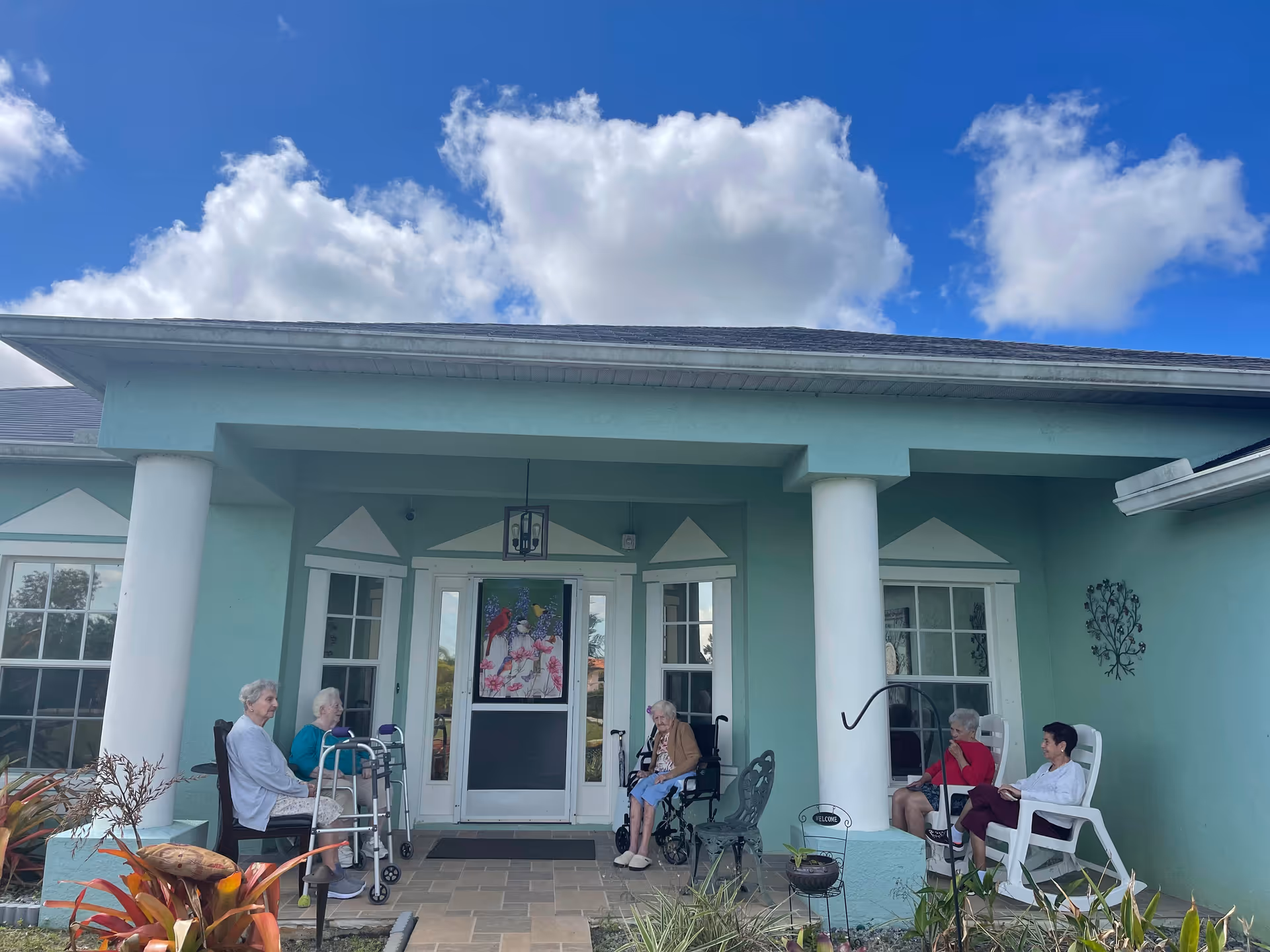 Five elderly individuals sitting and chatting on the front porch of a light blue building with white columns under a partly cloudy blue sky.