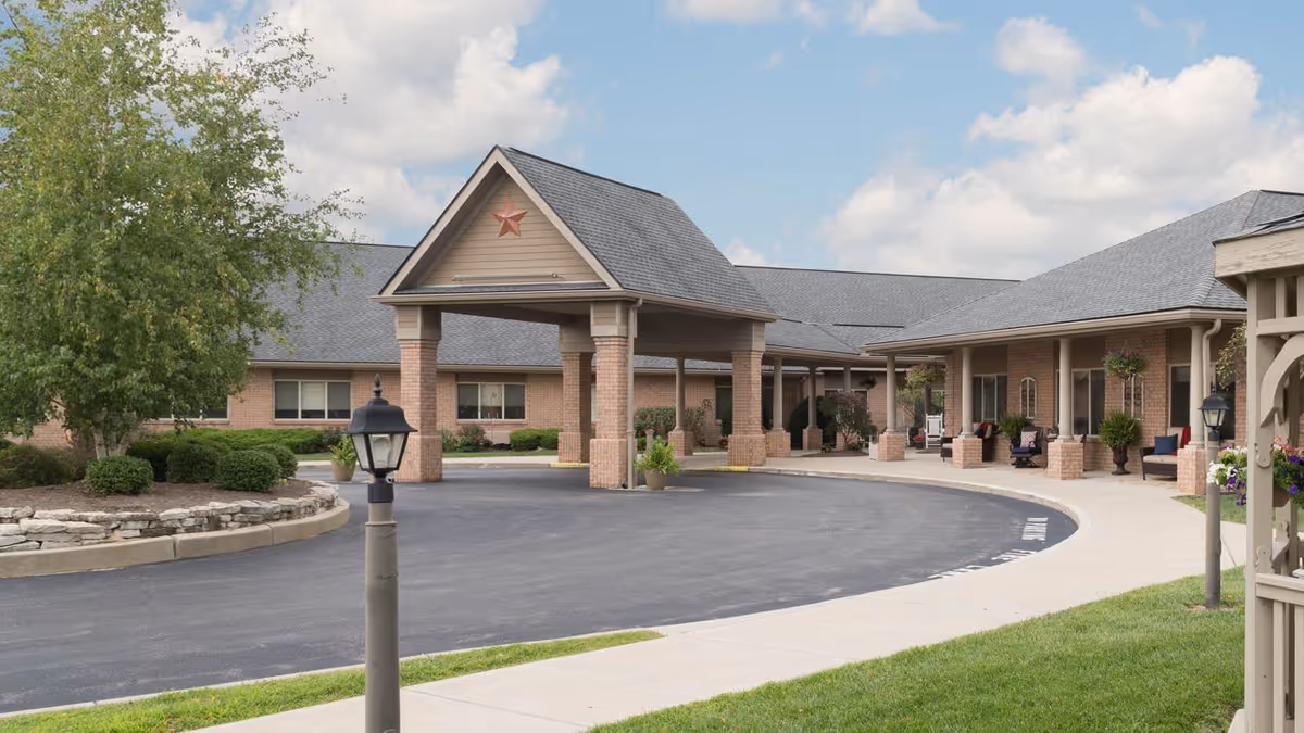 Front exterior view of a senior living facility with a covered entrance supported by brick columns, a circular driveway, landscaped greenery, and a partly cloudy sky.