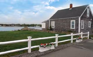 Small gray wooden building by water with a white fence and flower pots along the road.