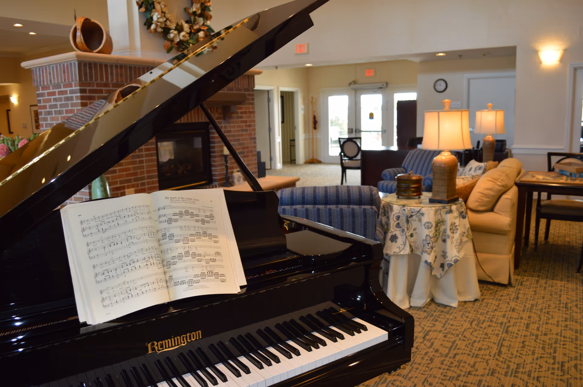 Grand piano with open sheet music in a cozy senior living lounge area furnished with sofas, lamps, and a fireplace.